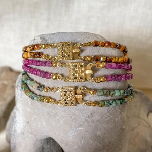 May include: Three bracelets stacked vertically, each featuring small faceted beads in amber, magenta, and teal. Each bracelet includes a central rectangular gold-toned metal clasp with an intricate floral cutout pattern and small gold-toned spacer beads.
