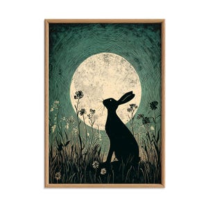 May include: Framed art print depicting a black hare silhouette observing a full moon. The artwork is set against a teal background with stylized grass and flowers, and is enclosed in a natural wood frame.