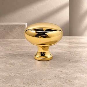 May include: A polished gold-tone cabinet knob featuring a smooth, rounded, mushroom-shaped head and a tapered cylindrical base. The metallic surface is highly reflective, showing light highlights and a warm, bright yellow-gold finish.