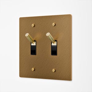 May include: A brass-colored, square light switch plate with two black toggle switches. The plate has a textured finish and is secured with four brass-colored screws. The toggle switches have cylindrical brass handles.