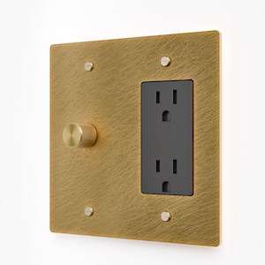 May include: A square wall plate with a brushed brass finish featuring a circular knurled knob on the left and a black dual electrical outlet on the right. The plate is secured by four flat-head screws, one at each corner of the metal faceplate.