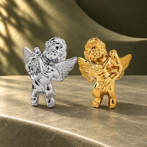 May include: Two cherub figurines, one with a polished silver finish and one with a polished gold finish. Each cherub has detailed wings, curly hair, and is holding a small bird. The figures are standing upright with a smooth, reflective surface.