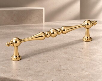 May include: A polished gold-tone metal cabinet pull featuring a central spherical accent flanked by tapered, elongated sections and rounded end caps. The handle is supported by two flared, cylindrical mounting posts at each end.