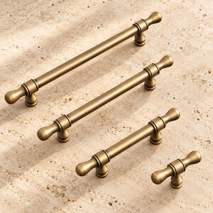 May include: Four metal cabinet pulls in a brushed antique brass finish, arranged in a diagonal line from longest to shortest. Each handle features a cylindrical bar, rounded end caps, and two circular mounting posts with a smooth texture.