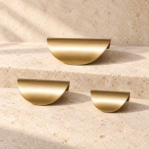 May include: Three semi-circular drawer pulls in a brushed brass finish. The hardware features a smooth, curved design with a flat top edge and a rounded bottom, presented in three different sizes arranged in a descending order.