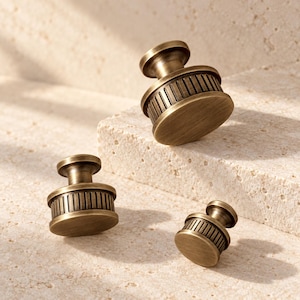May include: Three round cabinet knobs with a brushed antique brass finish. Each knob features a flat circular top, a central stem, and a cylindrical base with vertical ribbed detailing around the perimeter, creating a uniform appearance.