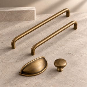 May include: A collection of four brass-toned metal cabinet hardware pieces, including two long arched bar pulls, one semi-circular cup pull, and one small round knob, all featuring a smooth, brushed metallic finish.