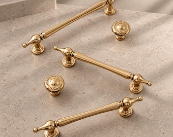 May include: A collection of gold-toned cabinet hardware including three elongated drawer pulls with tapered ends and two round knobs featuring a beaded edge detail. Each piece has a smooth, polished metallic finish and a classic, rounded shape.