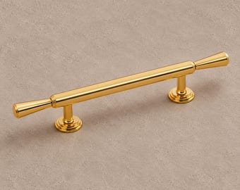 May include: A polished gold-tone metal cabinet pull featuring a long, cylindrical bar with tapered ends. The handle is supported by two circular, tiered mounting posts that connect to the main bar, creating a sleek and uniform metallic finish.