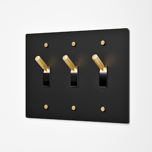 May include: A rectangular matte black wall plate featuring three toggle light switches. Each switch has a cylindrical gold-toned metal lever and a black base. The plate is secured with six small, round, gold-toned screws positioned at the top and bottom.