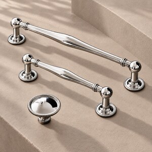 May include: Two polished silver-toned metal cabinet pulls with a smooth, tapered centre and spherical ends, accompanied by a single matching round cabinet knob. Each piece features a decorative beaded edge on the circular mounting bases.