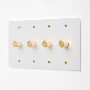 May include: A rectangular white wall plate featuring four cylindrical gold-toned knobs arranged horizontally in the center. The plate is secured by eight small, circular gold-toned screws, with four positioned along the top edge and four along the bottom edge.