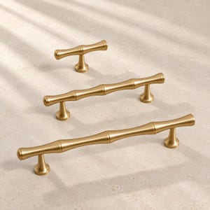Könnte beinhalten: Drei goldfarbene Metall-Schrankgriffe mit einem segmentierten, von Bambus inspirierten Design. Das Set enthält einen kleinen T-förmigen Knopf und zwei längere Griffe, die jeweils eine glatte, matte Oberfläche und ausgestellte, runde Sockel aufweisen.