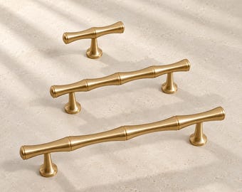 Op de afbeelding: Drie goudkleurige metalen kastgrepen met een gesegmenteerd ontwerp geïnspireerd op bamboe. De set bevat één kleine T-vormige knop en twee langere handgrepen, elk met een gladde matte afwerking en uitlopende ronde montagevoeten.