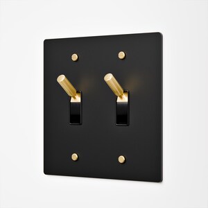 May include: A square matte black light switch plate featuring two toggle switches with metallic gold levers. The plate is secured by four matching gold-toned screws positioned at the top and bottom corners of each switch mechanism.
