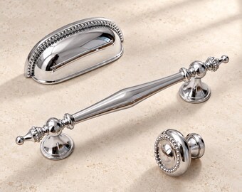 May include: A set of three polished chrome cabinet hardware pieces including a semi-circular cup pull with beaded edges, a long slender handle with decorative finials and central tapering, and a round knob featuring a beaded border design.