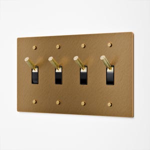 May include: A rectangular, brushed brass light switch plate with four black toggle switches and brass screws. The toggle switches have brass handles. The plate is mounted on a white wall.