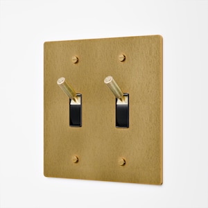 May include: A brass-colored, square light switch plate with two black toggle switches. The plate has a textured finish and is secured with four screws. The toggle switches have cylindrical handles.