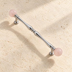 May include: A cabinet pull handle featuring a polished chrome finish with a slender, contoured metal bar. The handle is capped at both ends with smooth, spherical pale pink stone knobs, creating a balanced and symmetrical hardware piece.