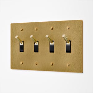 May include: A rectangular brass-coloured light switch plate with four black toggle switches. The plate has a textured surface and is mounted with small screws. The toggle switches have brass-coloured levers.