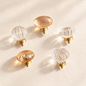 May include: Five cabinet knobs featuring clear and amber-tinted glass tops with gold-toned metal bases. Three knobs have a smooth, rounded surface, while two knobs feature a ribbed, fluted texture. Each knob is attached to a circular metal mount.