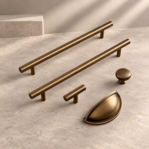 May include: A collection of antique brass cabinet hardware including two long bar pulls, one small T-bar knob, one round mushroom knob, and one semi-circular cup pull. Each piece features a smooth, brushed metallic finish and cylindrical stems.