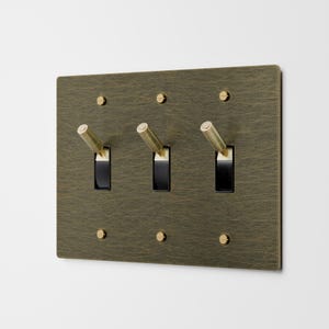 May include: A rectangular, brass-colored light switch plate with three black toggle switches and six small, round screws. The plate has a textured finish and is mounted on a white wall. The toggle switches have gold-colored handles.