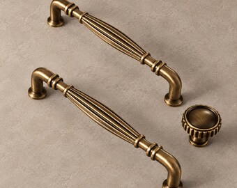 May include: Two antique brass cabinet pulls with a fluted, elongated center and curved ends, accompanied by a single matching round cabinet knob featuring a ridged edge and a smooth, concave center, all displaying a warm, metallic finish.