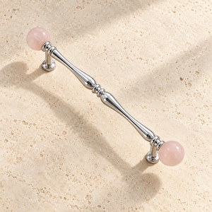 May include: A cabinet pull handle featuring a polished chrome finish with a slender, contoured metal bar. The handle is capped at both ends with smooth, spherical pale pink stone knobs, creating a balanced and symmetrical hardware piece.