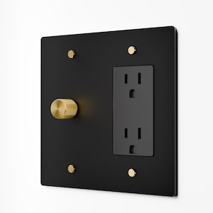 May include: A square matte black wall plate featuring a dual electrical outlet and a circular gold-toned knurled rotary dimmer knob. The plate is secured to the wall with four small, flat-head gold-toned screws positioned at each corner.