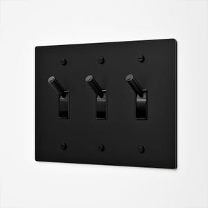 May include: A matte black triple toggle light switch plate featuring three cylindrical toggles and six matching black screws. The rectangular wall plate has a clean, flat surface with a uniform dark finish and a modern, industrial aesthetic.