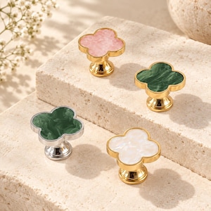 May include: Four clover-shaped cabinet knobs with smooth, marbled-effect inserts. Two knobs feature gold-tone metal bases with pink and white inserts, while two feature green inserts with one set in a gold-tone base and one in a silver-tone base.