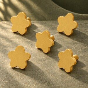 May include: A set of five gold-toned metal cabinet knobs, each shaped like a five-petaled flower with a smooth, matte finish. The knobs are arranged in a scattered pattern, showing the front-facing floral design and the cylindrical base.
