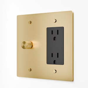 May include: A square wall plate with a brushed gold finish featuring a black electrical socket and a circular gold dimmer knob with a textured knurled edge. The plate is secured to the wall with four small gold screws at the corners.