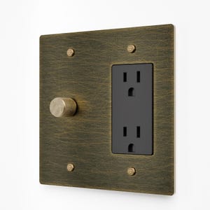 May include: A square wall plate with a brushed antique brass finish featuring a textured, scratched surface. It includes a black dual electrical outlet and a single circular, knurled brass knob. The plate is secured by four matching brass screws.