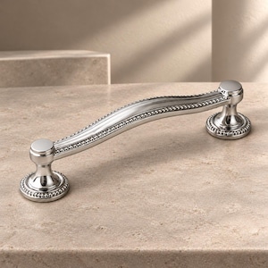 May include: A polished silver-toned metal cabinet pull with a curved, arched handle. The design features a central smooth bar flanked by beaded detailing along the edges, terminating in two circular bases with matching beaded rims.
