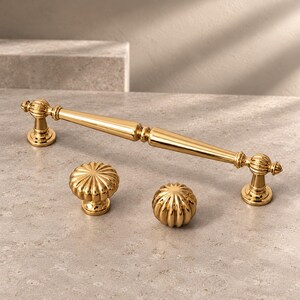 May include: A polished gold-tone metal cabinet handle with a long, tapered center and two round, fluted knobs. The handle features decorative spherical accents at the mounting points, while the two separate knobs have a distinct ribbed design.