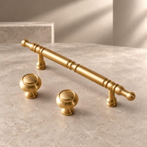 May include: A set of hardware consisting of one long, cylindrical cabinet pull with a decorative turned profile and two matching round cabinet knobs, all finished in a smooth, brushed satin gold metal tone.