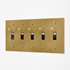 May include: A rectangular brass light switch plate with five toggle switches. The plate has a textured finish and is mounted with small screws. The toggle switches are gold-colored and have black switch openings.