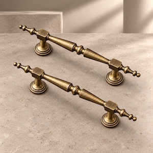 May include: Two antique brass cabinet pulls featuring a tapered, spindle-like design with square mounting blocks and circular bases. The metal hardware has a brushed, weathered finish with visible texture and pointed finial ends.