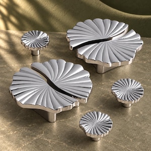 May include: A collection of five silver-toned metal cabinet knobs and pulls featuring a reflective, fan-shaped design with radiating grooves. The set includes two larger, wide fan-shaped pulls and three smaller, circular knobs with matching radial textures.