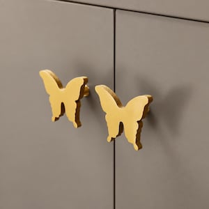 Könnte beinhalten: Zwei goldfarbene schmetterlingsförmige Schrankknöpfe an einer einfarbigen grauen Schranktür. Die Knöpfe haben eine glatte, metallische Oberfläche und sind in der Silhouette eines Schmetterlings mit weit ausgebreiteten Flügeln geformt.