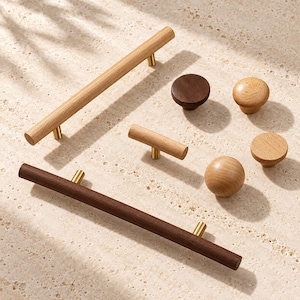 May include: A collection of wooden cabinet hardware including two long bar pulls, one short T-bar pull, and three round knobs. The hardware features light and dark wood finishes with gold-toned metal mounting posts for attachment to furniture.