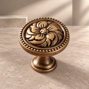 May include: A round cabinet knob with an antique brass finish featuring a central floral relief pattern. The circular face is bordered by a beaded edge, and the knob is supported by a tapered pedestal base with a matching metallic texture.