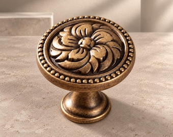 May include: A round cabinet knob with an antique brass finish featuring a central floral relief pattern. The circular face is bordered by a beaded edge, and the knob is supported by a tapered pedestal base with a matching metallic texture.