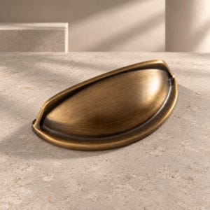 Könnte beinhalten: Ein halbkreisförmiger Muschelgriff für Schubladen mit gebürstetem Antikmessing-Finish. Der Beschlag verfügt über eine glatte, gewölbte Oberfläche mit einer vertieften Griffmulde und zwei Befestigungspunkten an der flachen Basis.