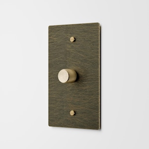 May include: A rectangular, dark brass dimmer switch plate with a textured surface. It features a round, textured knob in the center and two small screws. The plate is mounted on a white wall.