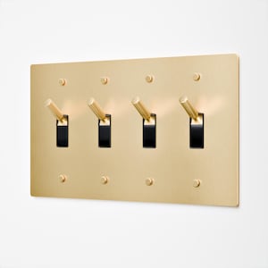 May include: A gold-coloured light switch plate featuring four black toggle switches. Each switch has a gold-coloured lever. The plate is mounted on a white wall. The plate is secured with eight small, gold-coloured screws.