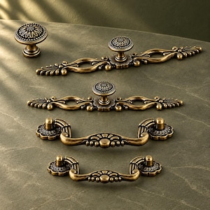 May include: A collection of five antique brass-toned metal cabinet hardware pieces, including one round knob, two long decorative pulls with central knobs, and two drop-style drawer pulls, all featuring intricate floral and scrollwork details.