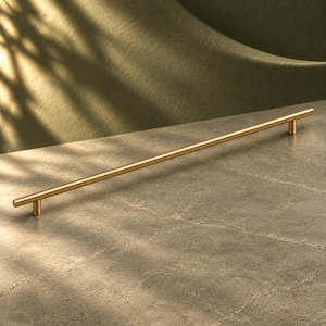 May include: A long, slender cabinet pull handle with a brushed gold finish. The hardware features a cylindrical bar design supported by two small, circular posts at each end, creating a clean and linear aesthetic for furniture or cabinetry.